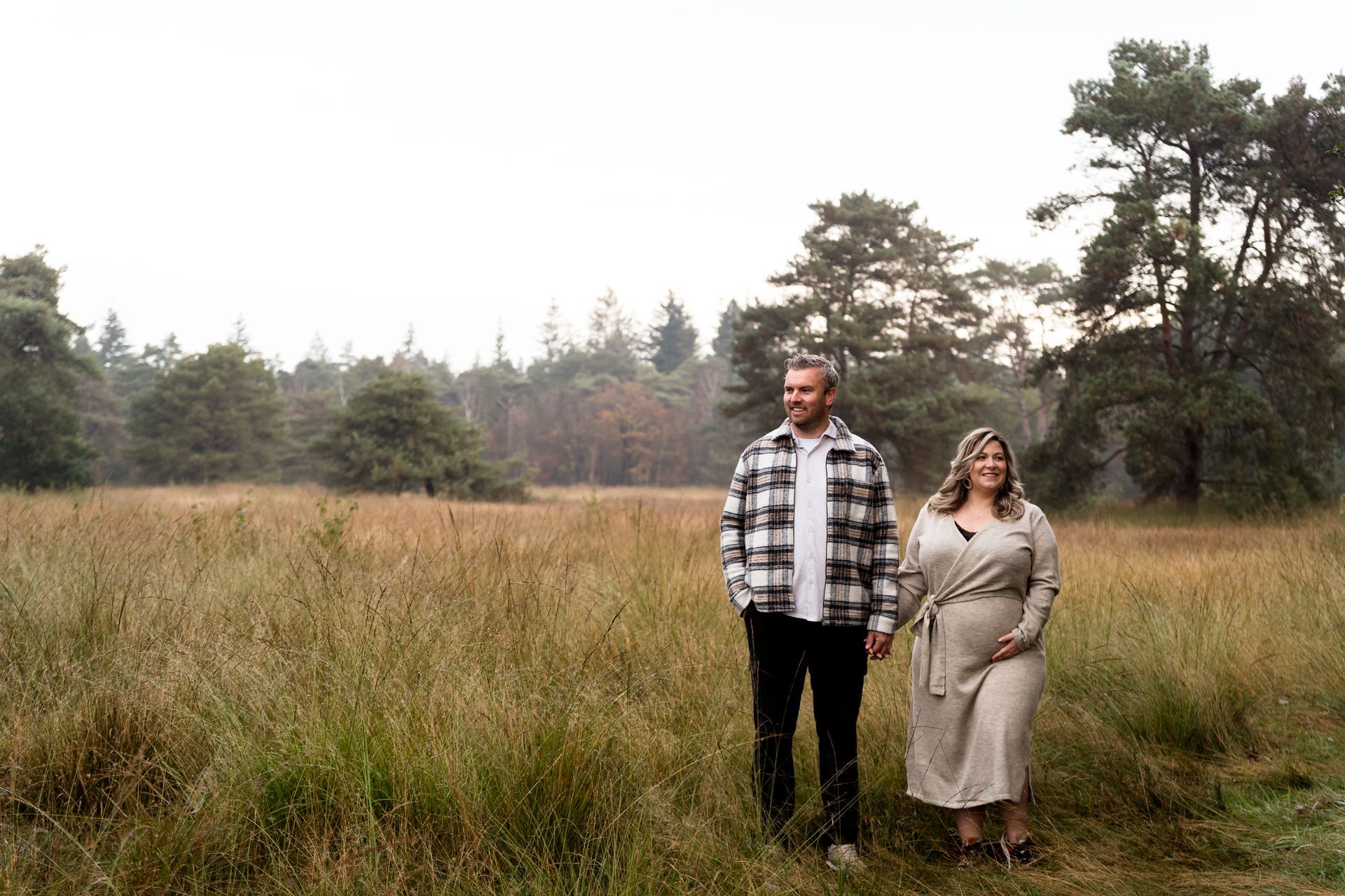 Een stel loopt hand in hand in een grasveld; de man draagt ​​een geruit overhemd, de vrouw een beige jurk. Op de achtergrond zijn bomen te zien.