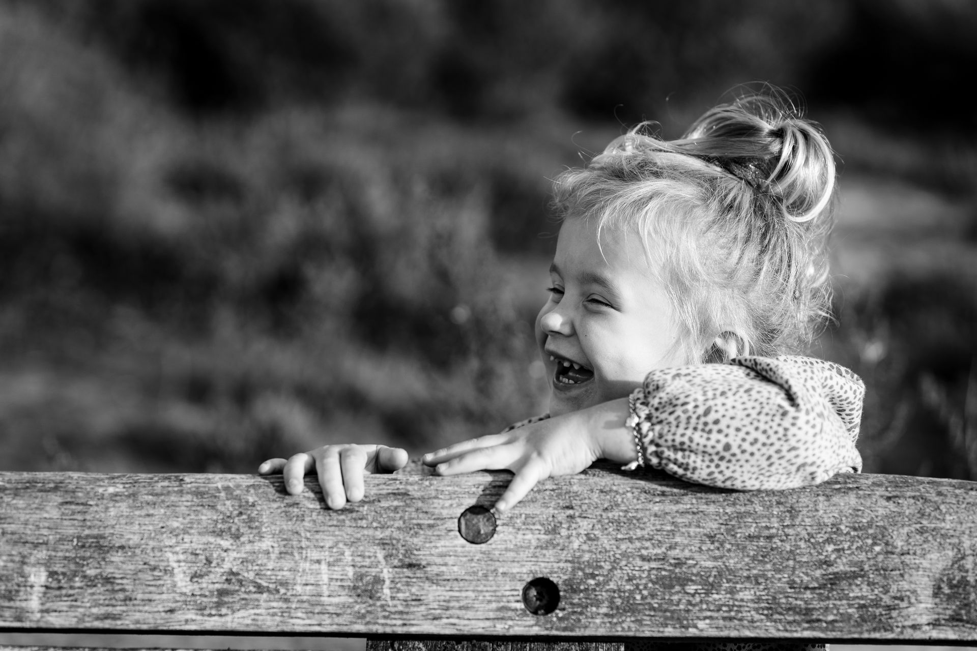 Jong kindje lacht en leunt buiten tegen een houten hek, met het haar in een knotje.