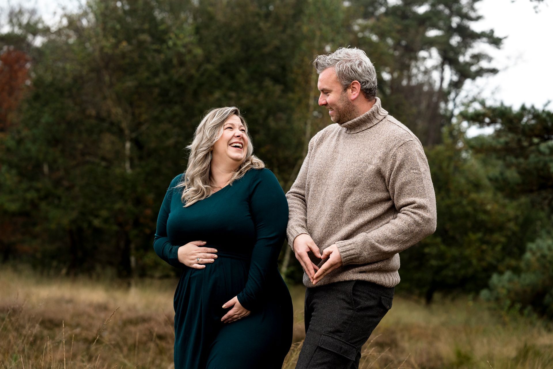 Een stel in de buitenlucht; de zwangere vrouw lacht, de man maakt een hartje met zijn handen en kijkt elkaar aan.