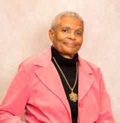 Elderly Black woman with short hair, smiling in a pink jacket and black turtleneck.