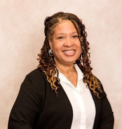 Woman with long, braided hair smiles, wearing a white shirt and black jacket. Neutral background.