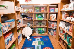 A children's bookstore with wooden shelves filled with toys and books, a blue hopscotch rug.