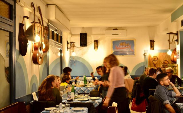 Persone che cenano in un ristorante illuminato da una luce calda, con quadri blu alle pareti e lampade a sospensione.
