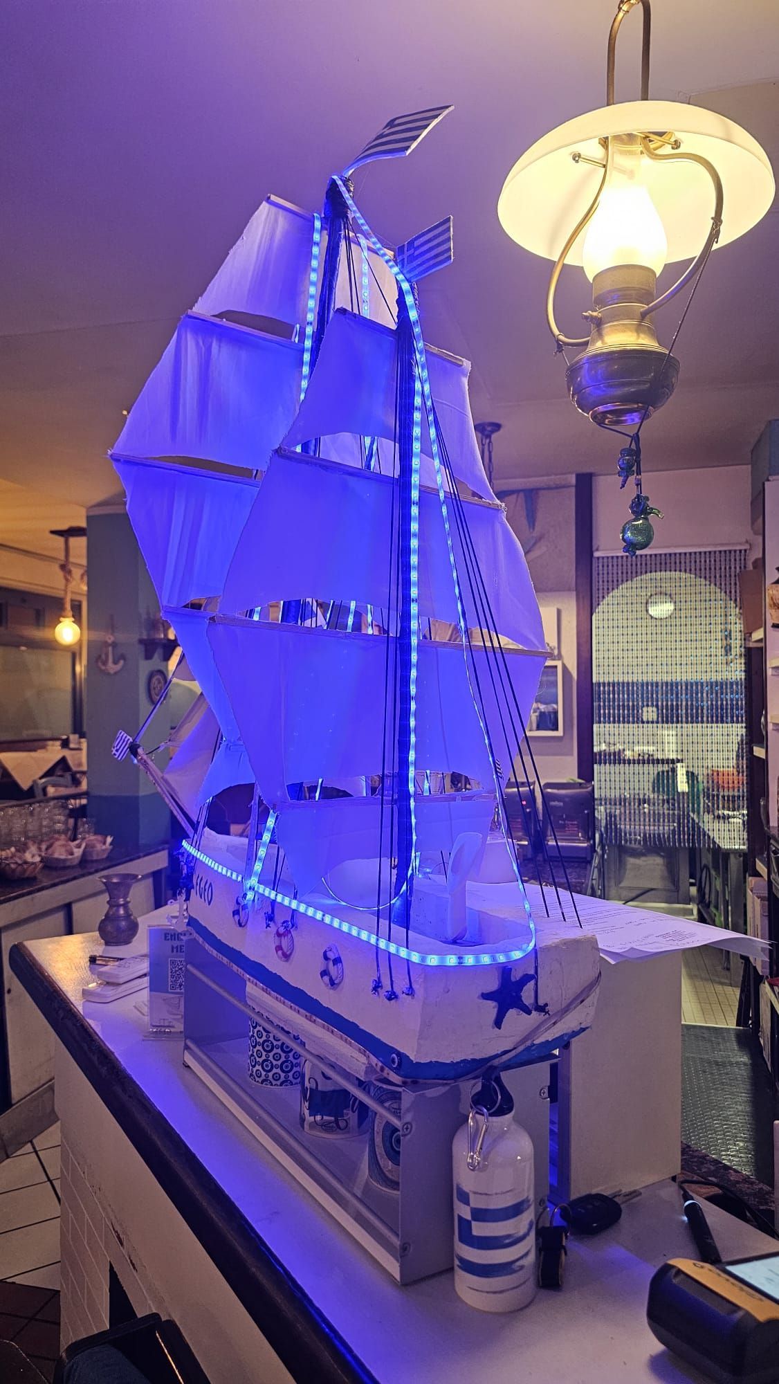 Esposizione al chiuso di un modellino di barca a vela illuminato di blu, con vele gonfiate dal vento e una lanterna accanto.