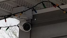White security camera mounted under an eave, next to Christmas lights.