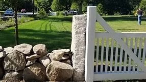 Stone wall and white gate opening onto a grassy lawn; a person walks in the background.