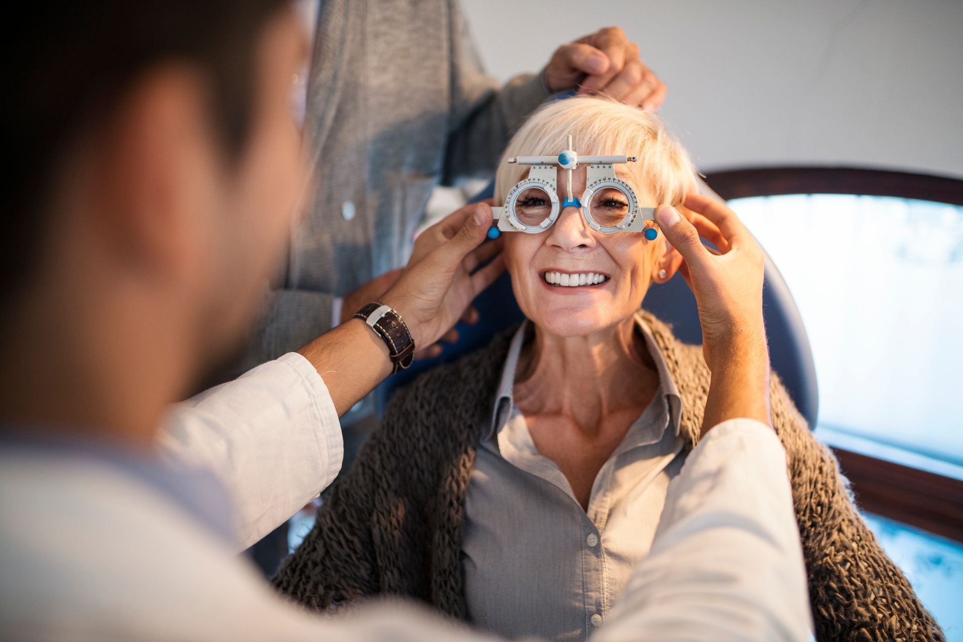 Optometrist fitting trial lenses on a smiling patient in an exam room.