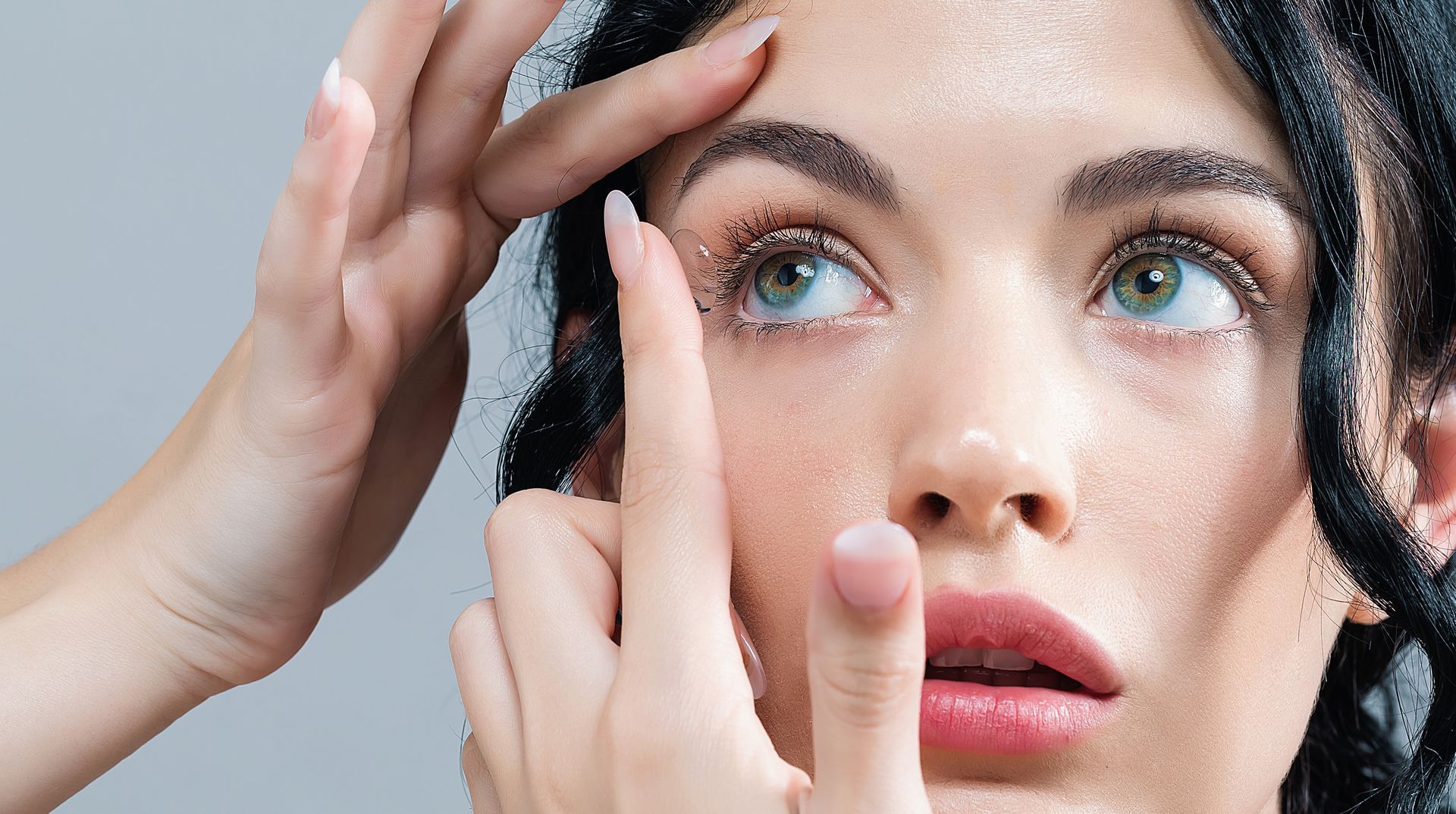 Woman putting in a contact lens, looking up. One hand holds her eyelid open; her other hand holds the lens.