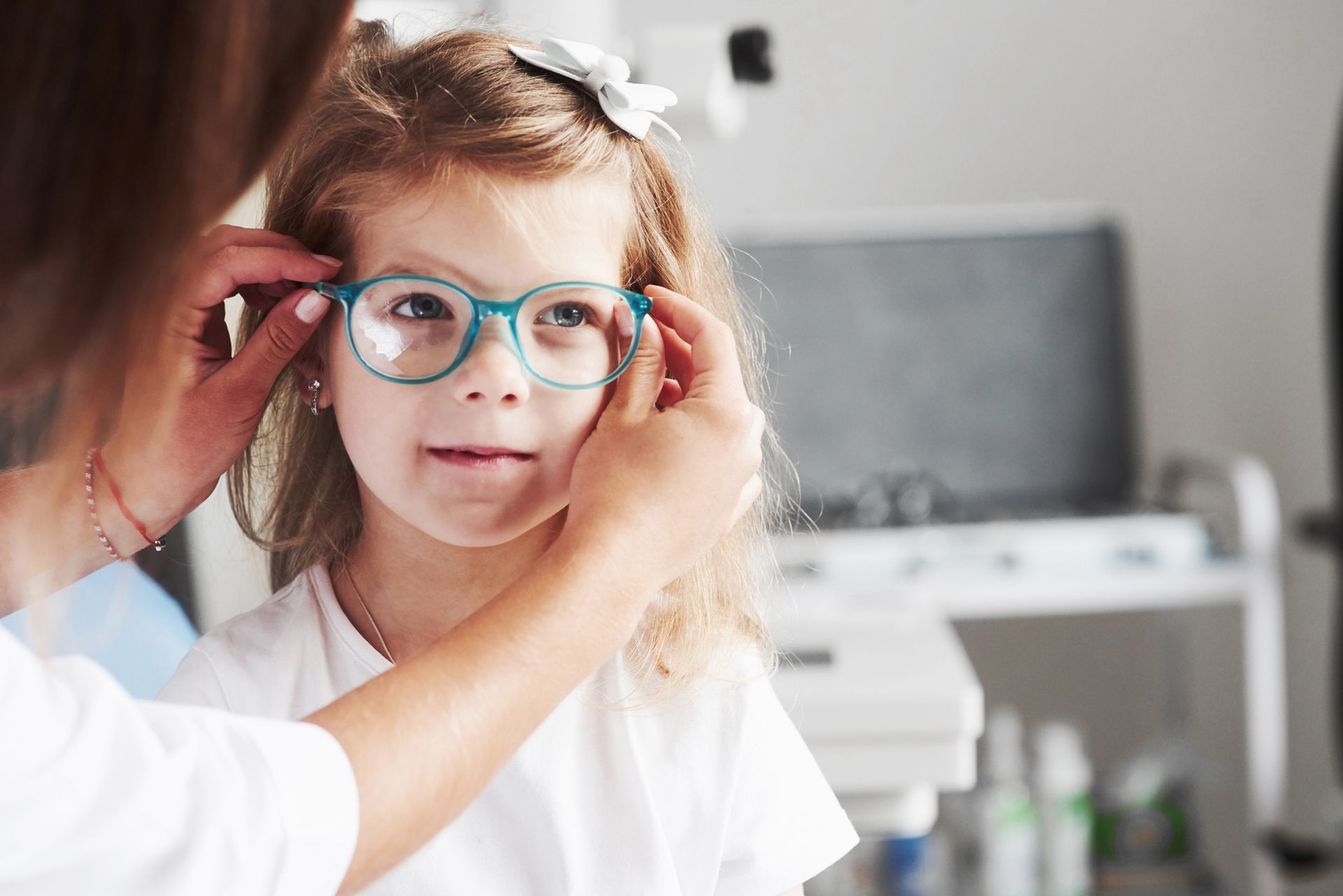 A person adjusting eyeglasses on a child. The child smiles in an eye exam setting.