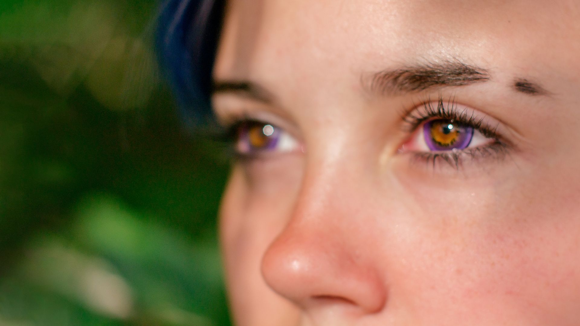 Close-up of person with purple eyes, nose, and eyebrow piercing; blurred green background.