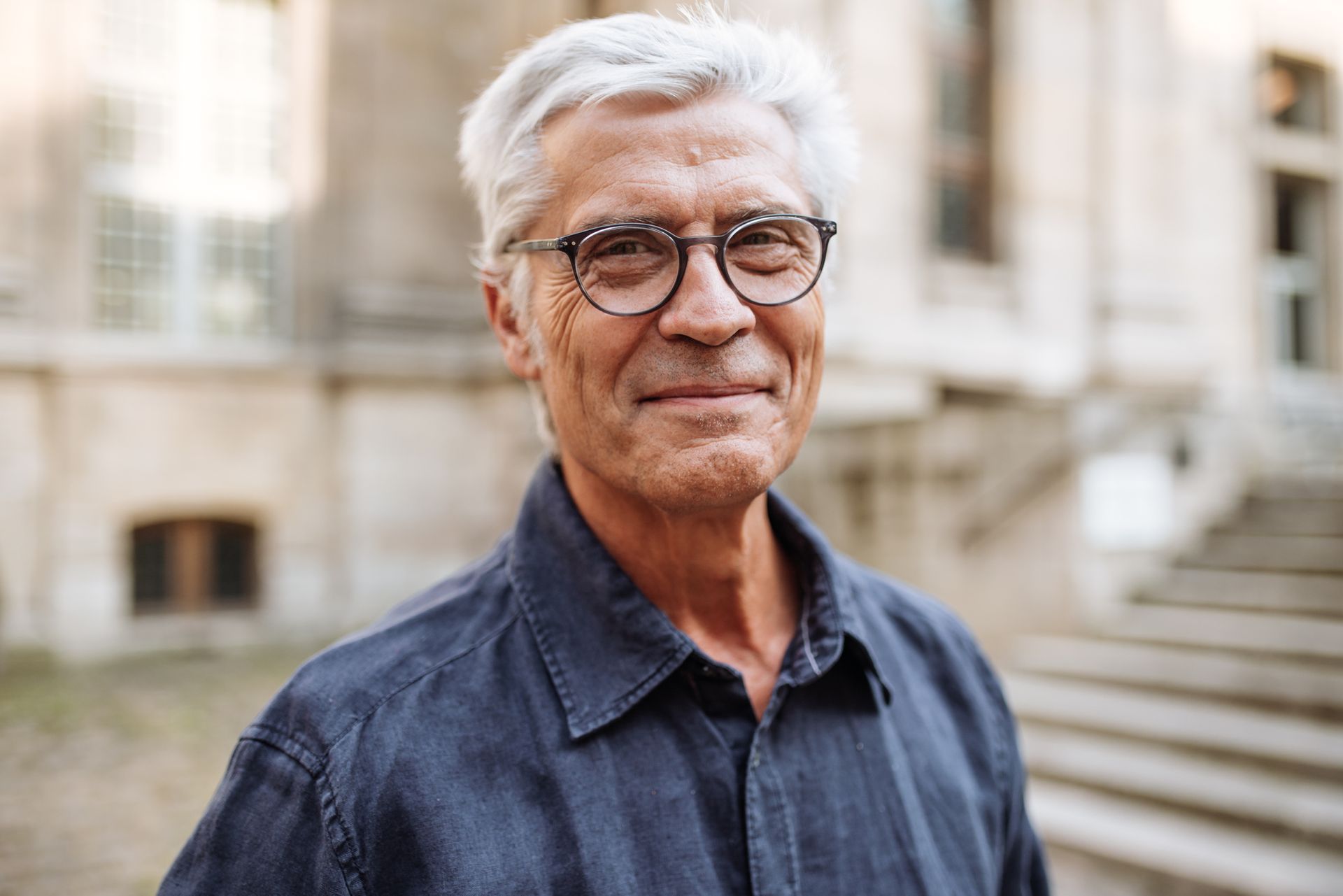 Man with gray hair and glasses smiles in front of a stone building.