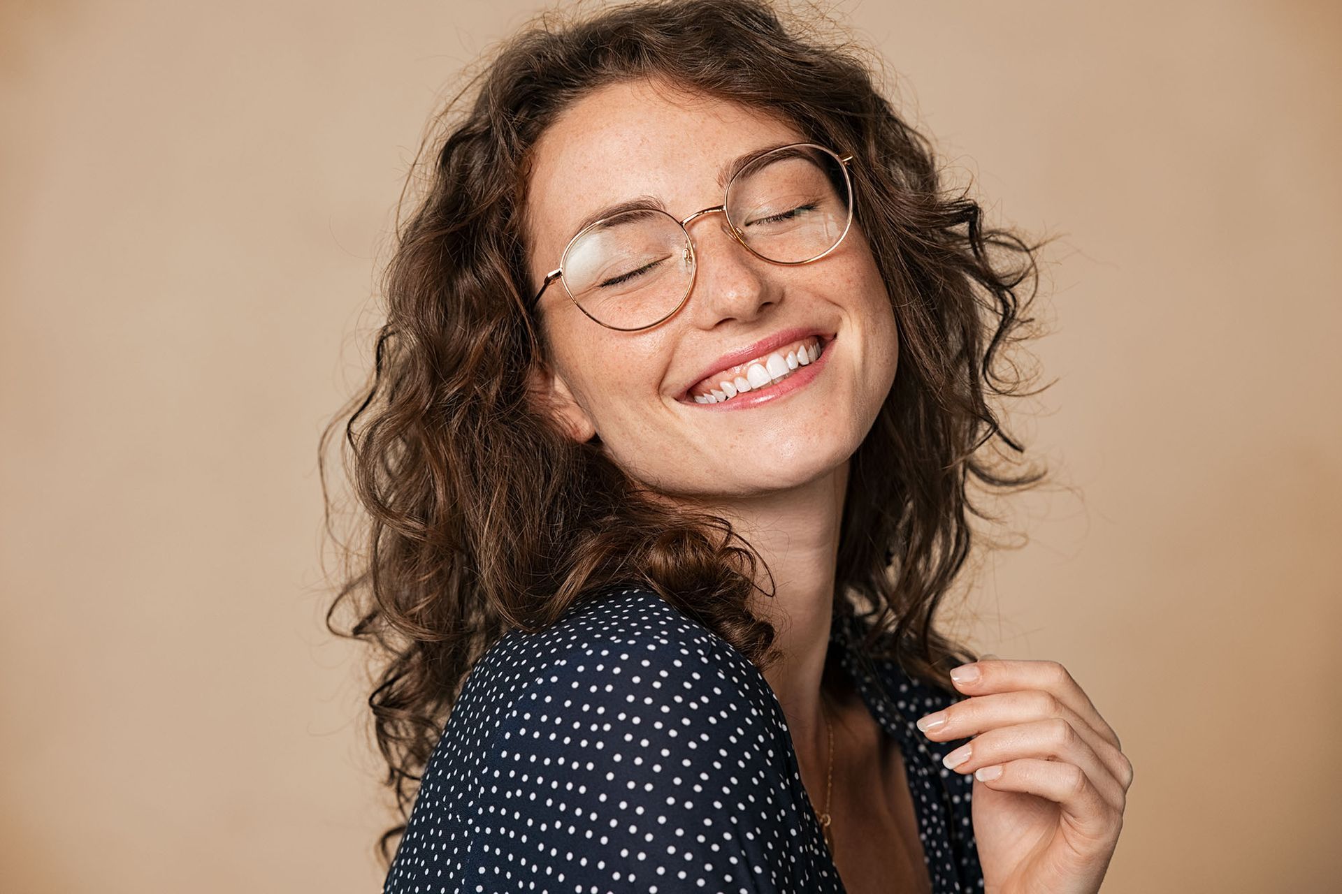 Woman with curly hair smiles, wearing glasses, against a beige background.
