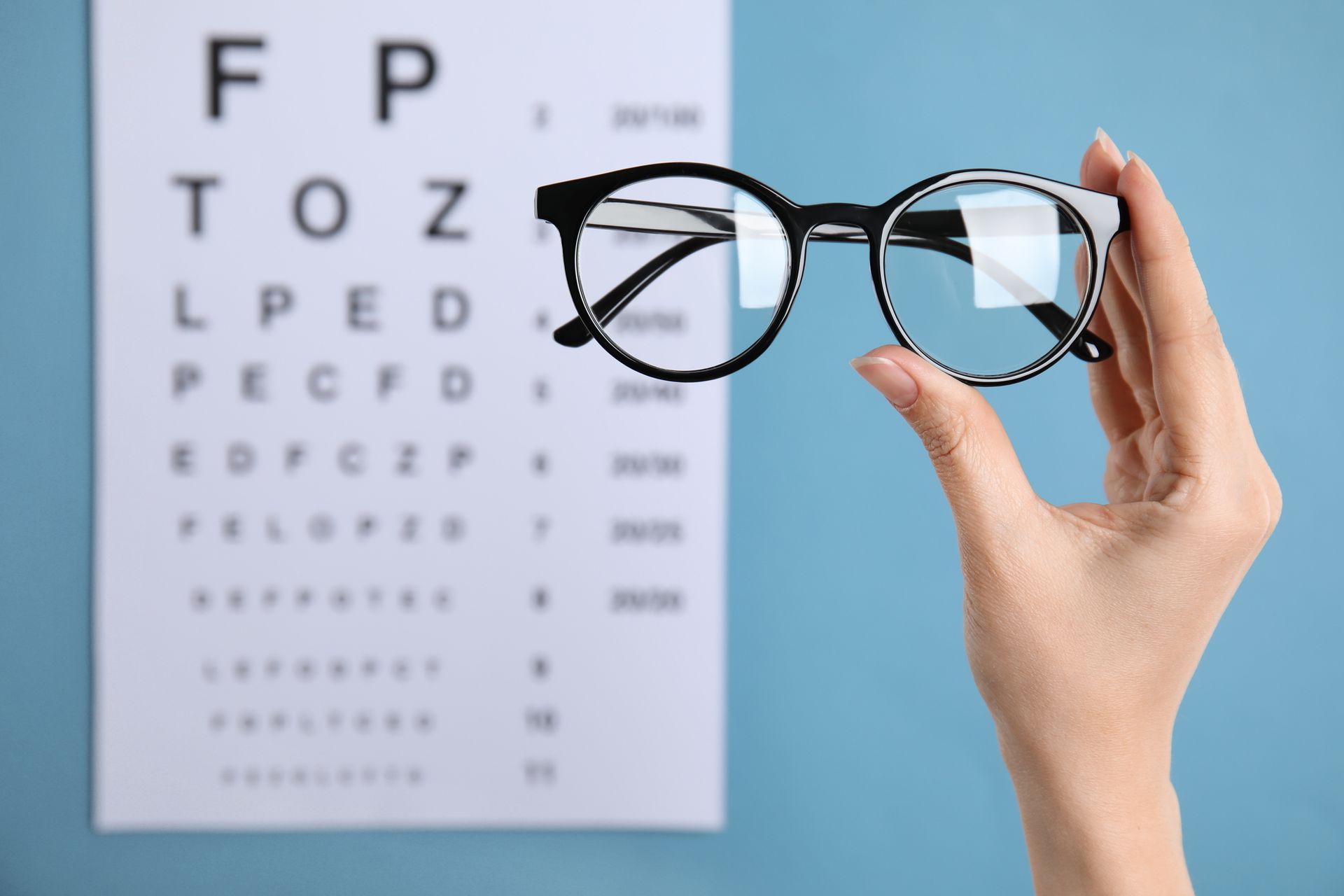 Hand holding eyeglasses in front of a blurred eye chart, blue background.