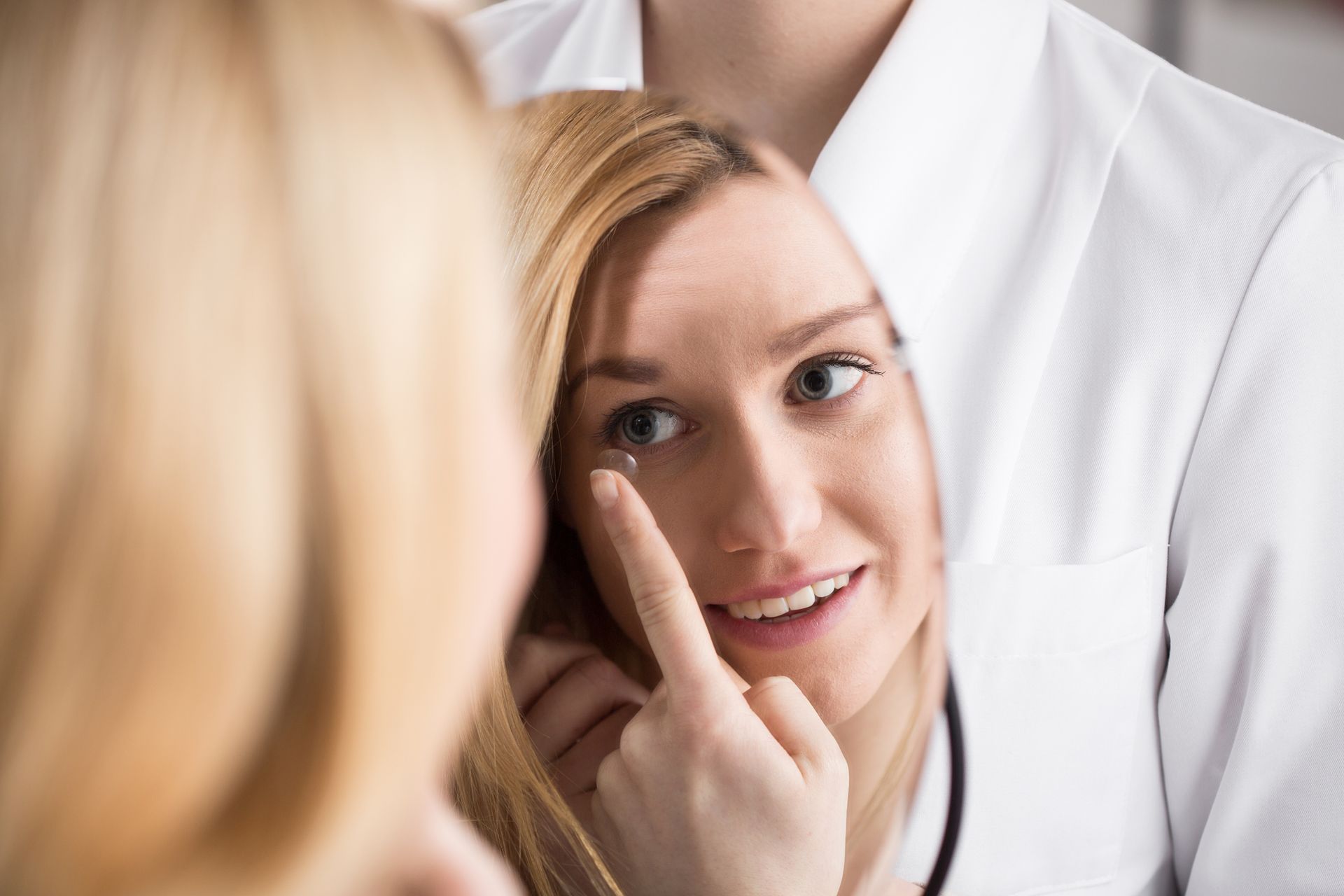 Woman inserting a contact lens in front of a mirror, being assisted by someone in a white coat.