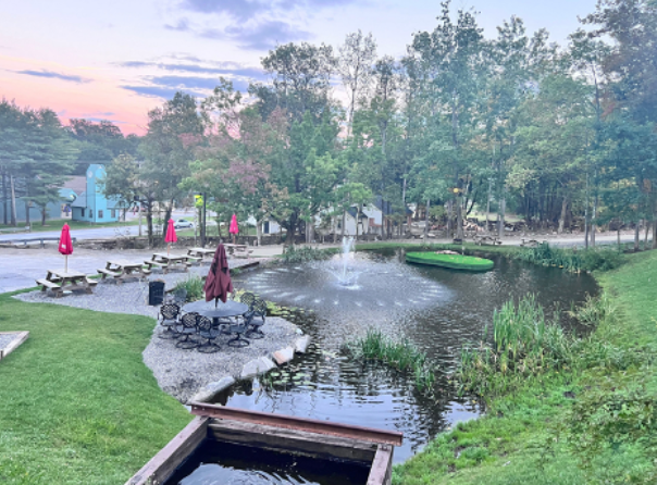 A pond with a fountain, surrounded by greenery and a seating area with tables and umbrellas.