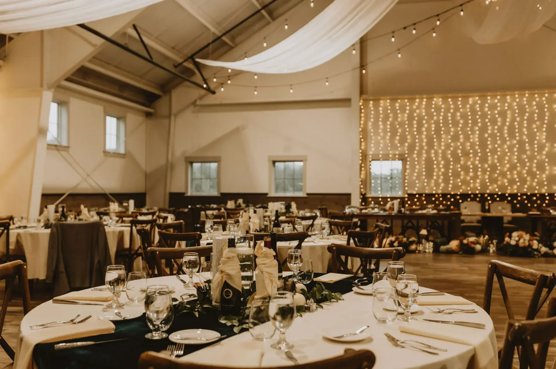 Reception hall decorated for an event with round tables, string lights, and draping fabric.