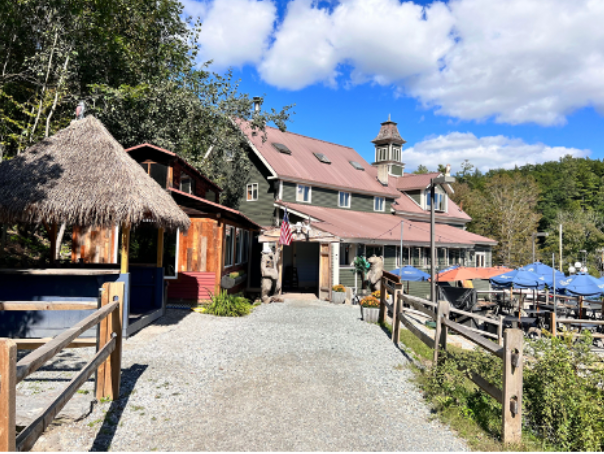 Stone path leads to a large, green building with a smaller hut beside it, under a bright blue sky.