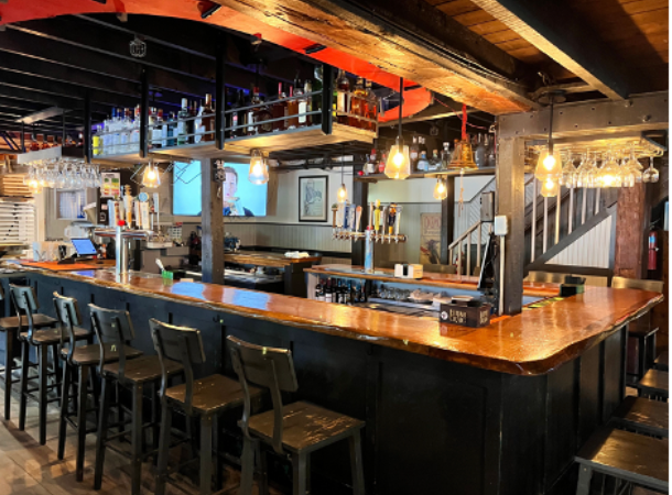 Interior bar with wooden counter, stools, and shelves stocked with bottles.