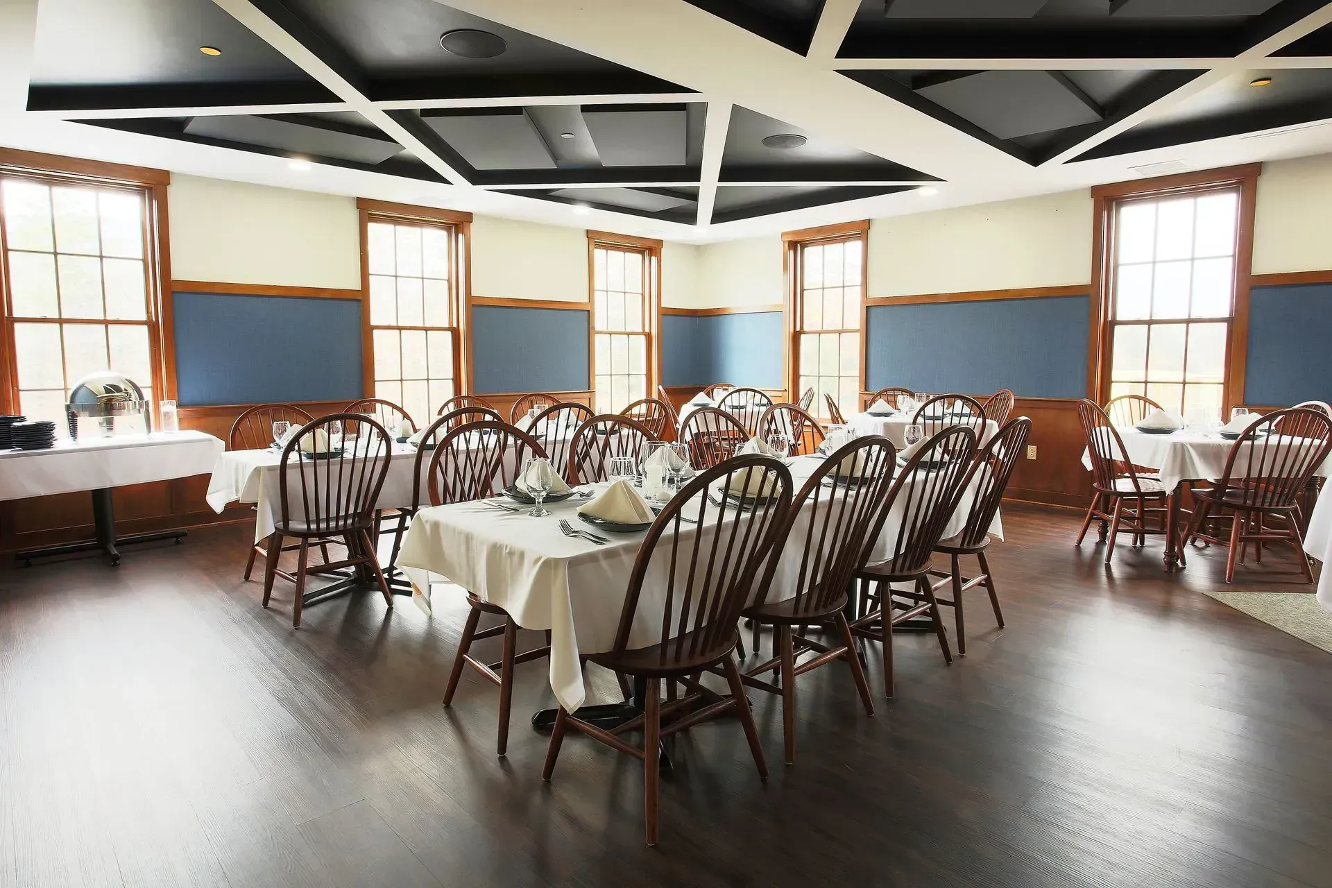 Dining room with wood tables, chairs, and windows. White tablecloths, blue walls, dark ceiling.