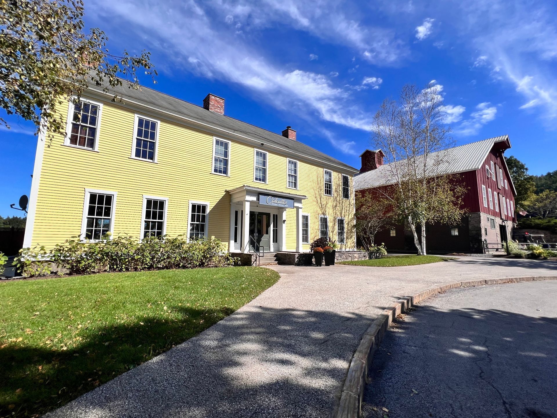Yellow farmhouse and red barn under a blue sky with clouds. Driveway leads to entrance.