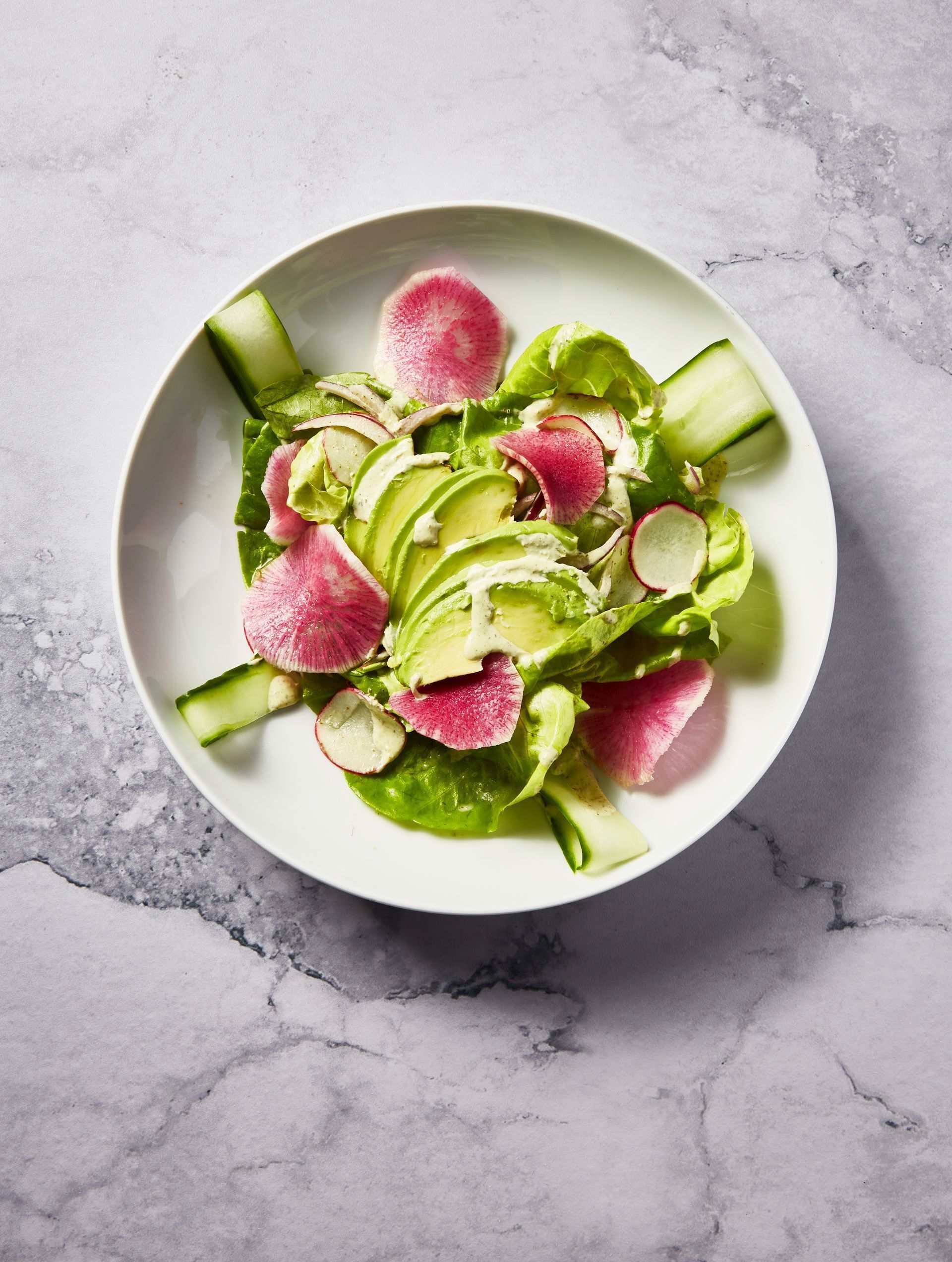 Salad with avocado, radishes, cucumbers, and lettuce on a white plate; marble background.