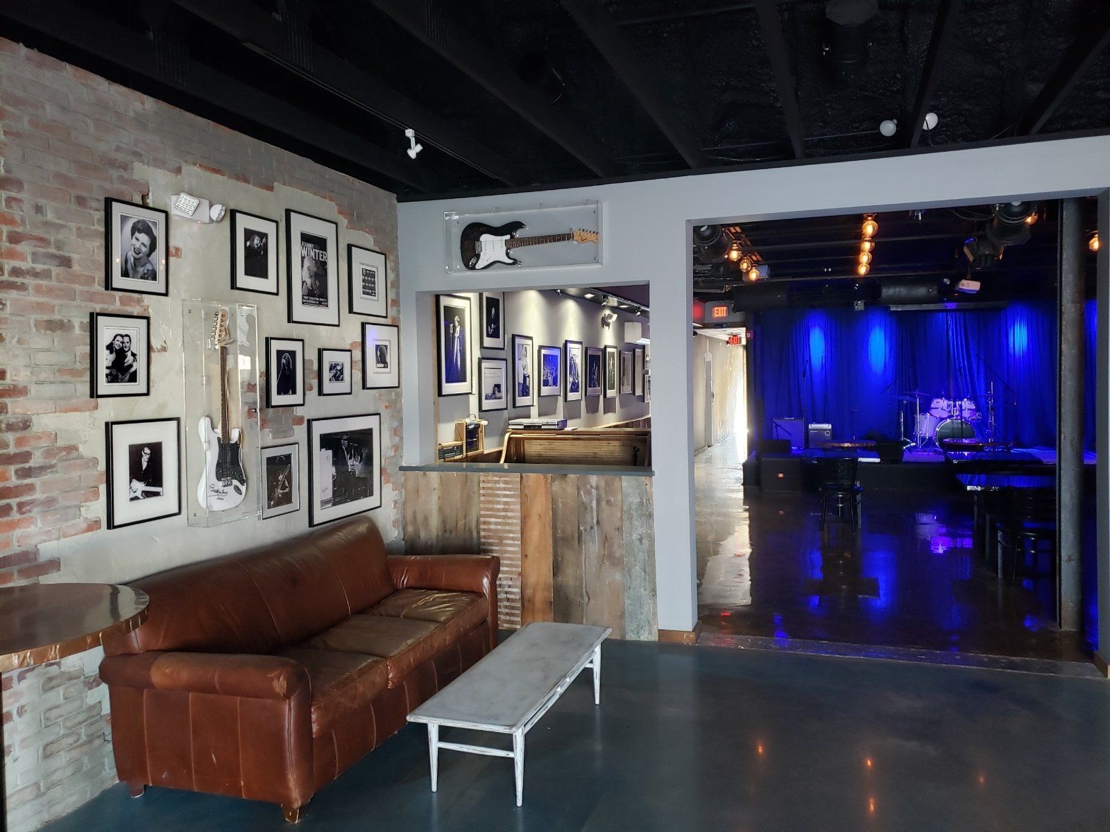 Lounge area with exposed brick, leather couch, and framed photos leading to a stage lit with blue lights.