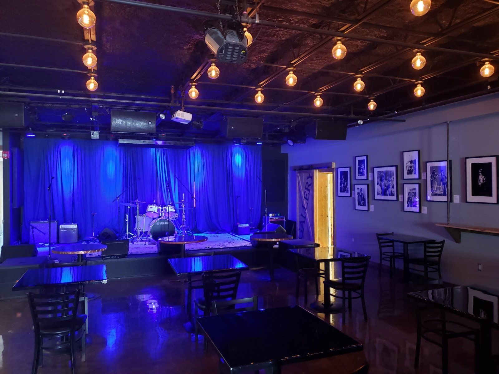 An empty music venue stage bathed in blue light; tables and chairs in the foreground, photos on the wall.