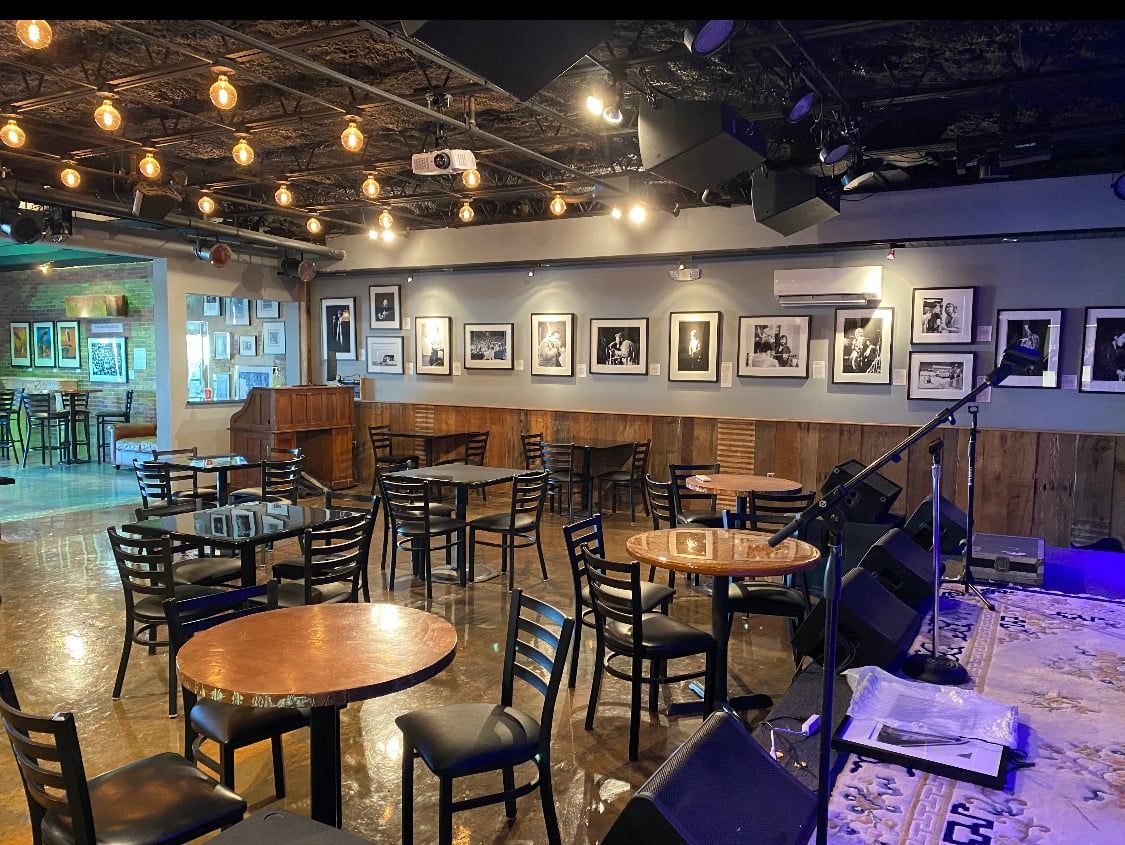 Interior of a music venue with tables, chairs, stage, photos on the wall, and string lights.