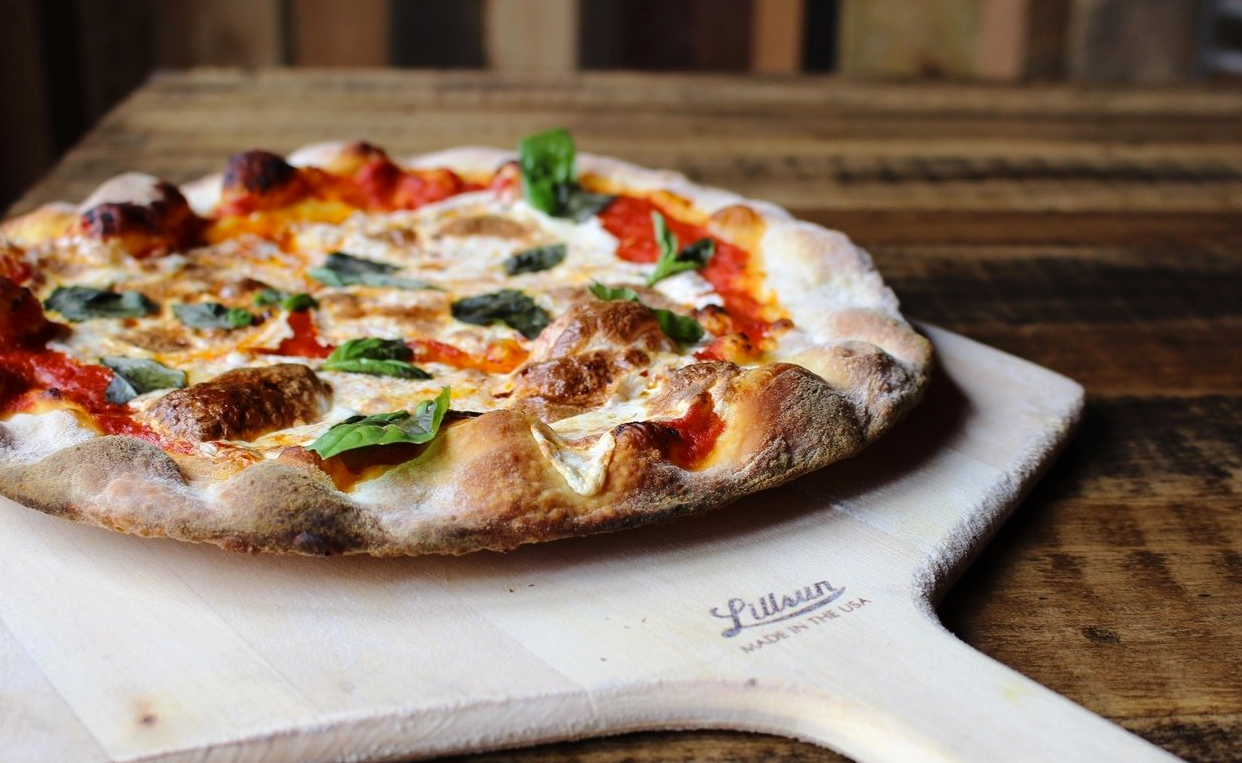 Pizza on a wooden peel, set against a wood background. Pizza has basil and tomato sauce.