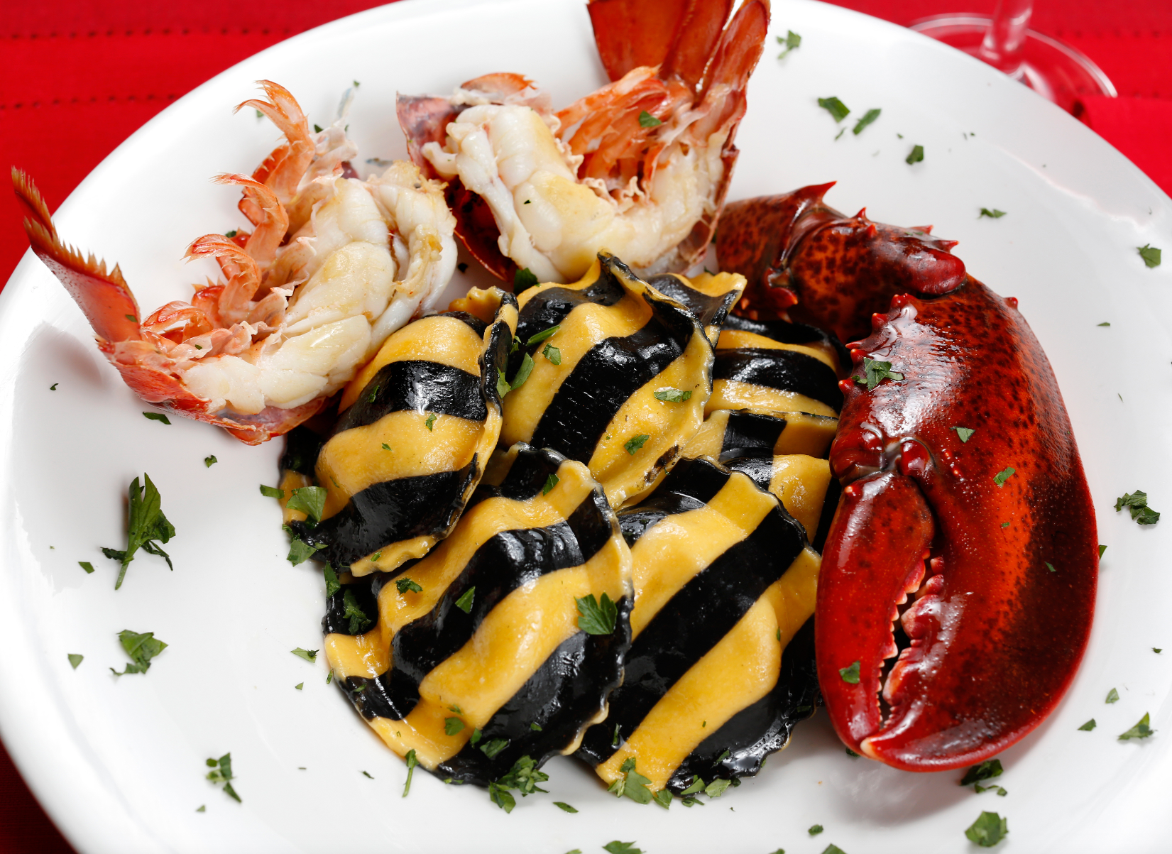 Lobster ravioli on a white plate with lobster tail and claw garnish. Black and yellow pasta with parsley.