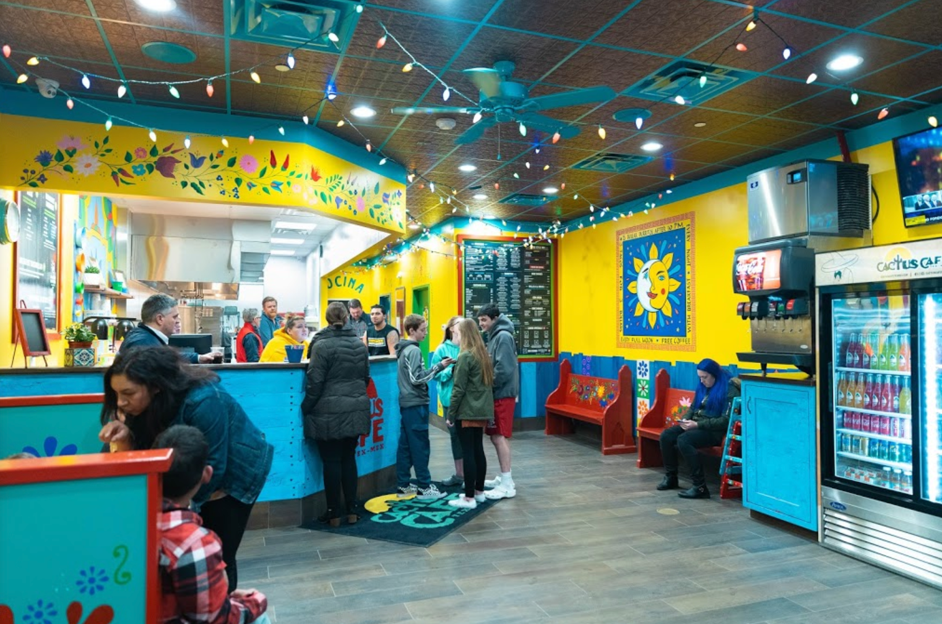 Vibrant interior of a Mexican restaurant. Customers line up near the counter; colorful decor with bright blue, yellow, and red accents.