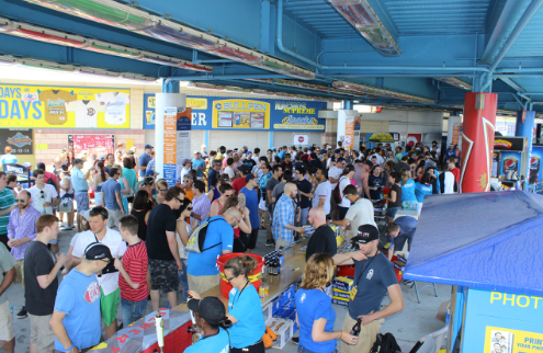 Crowd of people under a blue-painted structure at an outdoor event, some at vendor tables, many socializing.