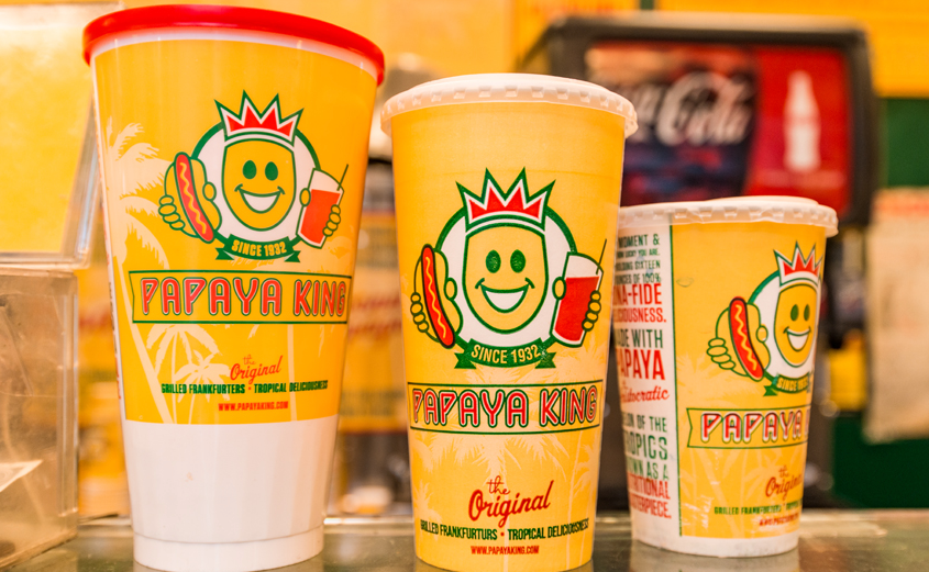 Three Papaya King cups of varying sizes, orange with the logo, sit on a counter.