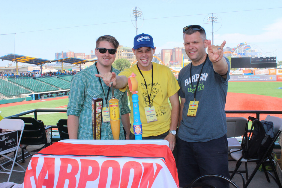 Three men at a baseball game booth. They hold up novelty beer tap handles, with a city skyline in the background.