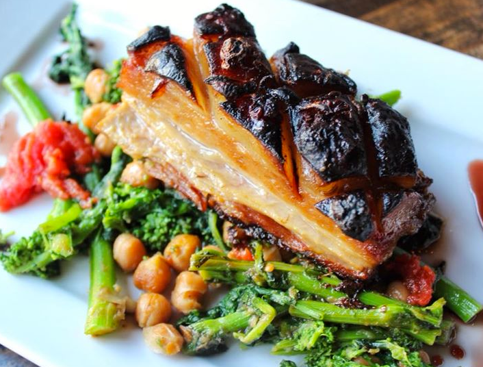 Roasted pork belly on bed of greens, chickpeas, and a red garnish, served on a white plate.