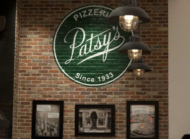 Patsy's Pizzeria sign on brick wall, above framed photos. Black pendant lights illuminate the sign.