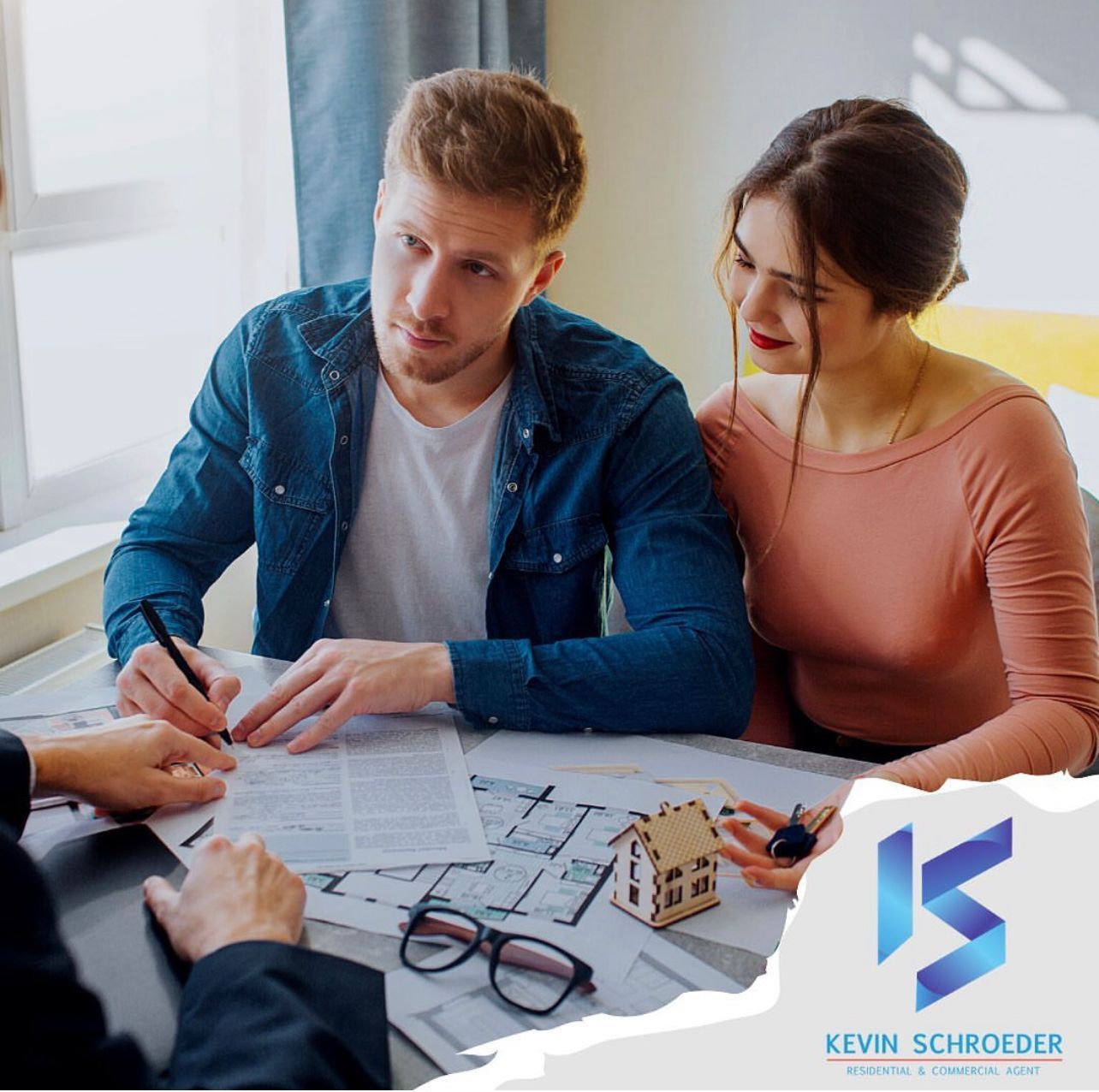 Couple signing documents with a realtor, blueprints, and house model on the table.