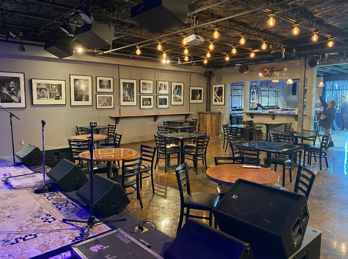 Empty music venue with stage, tables, chairs, and bar in background.