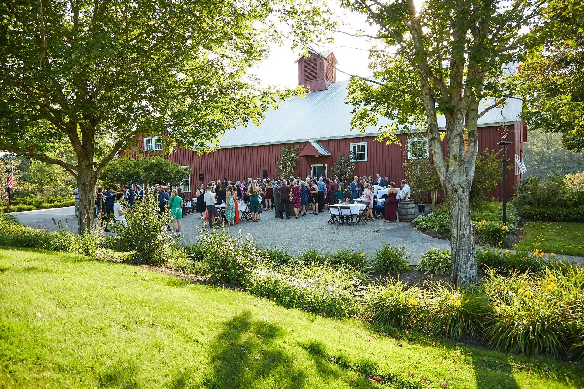Large outdoor gathering in front of a red barn. People mingle on a gravel patio and lawn, trees surround the scene.