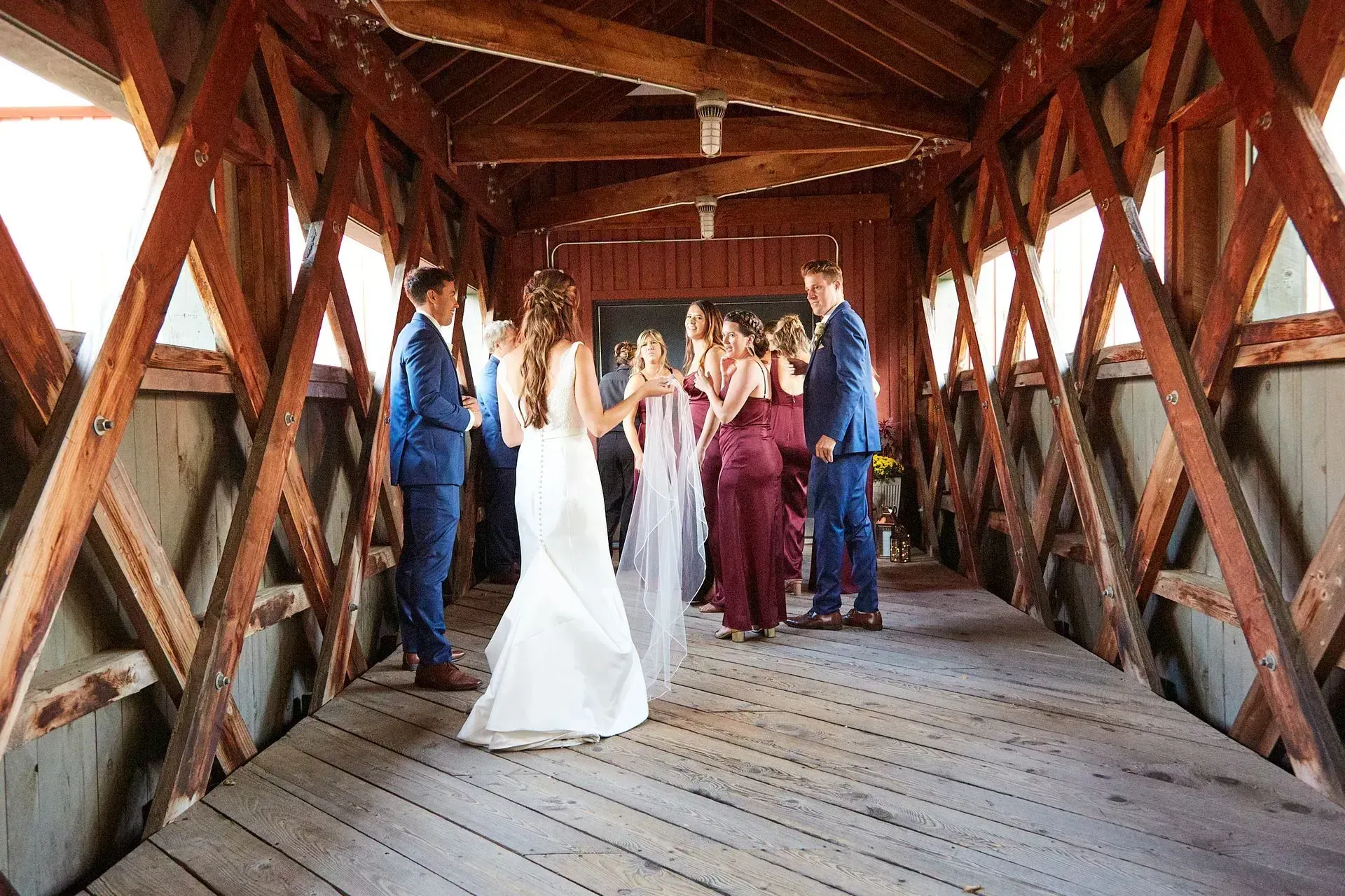 Wedding party inside a wooden covered bridge; bride in white dress, groom in blue suit, bridesmaids in burgundy dresses.