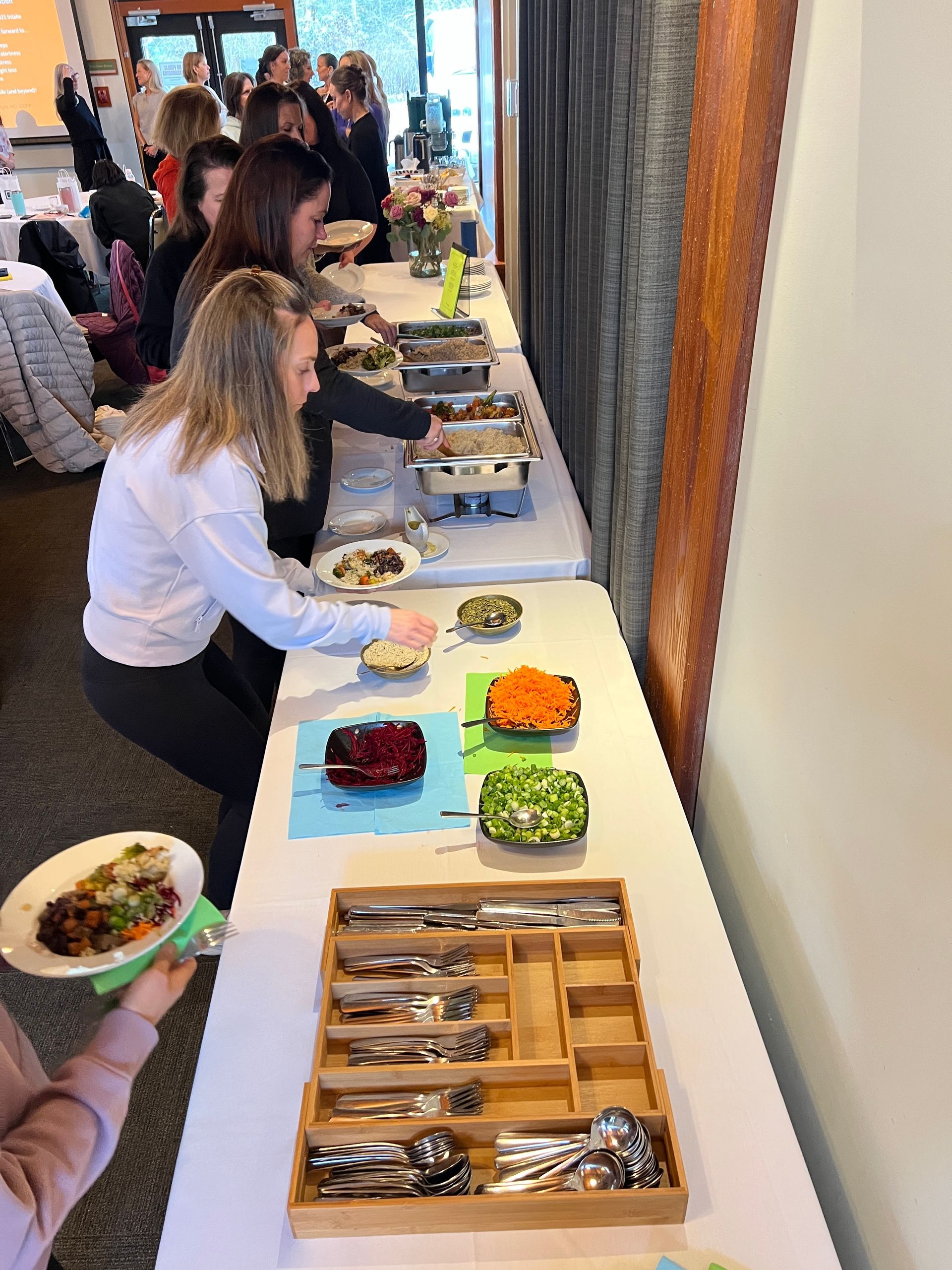 People serving themselves at a buffet. Several dishes of food are laid out along a white tablecloth.  A utensil organizer is visible.