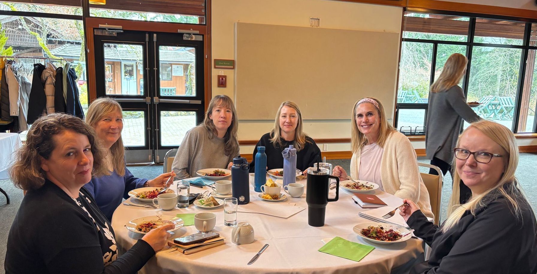 A group of seven women seated around a round table indoors, possibly at a luncheon or meeting, smiling at the camera.
