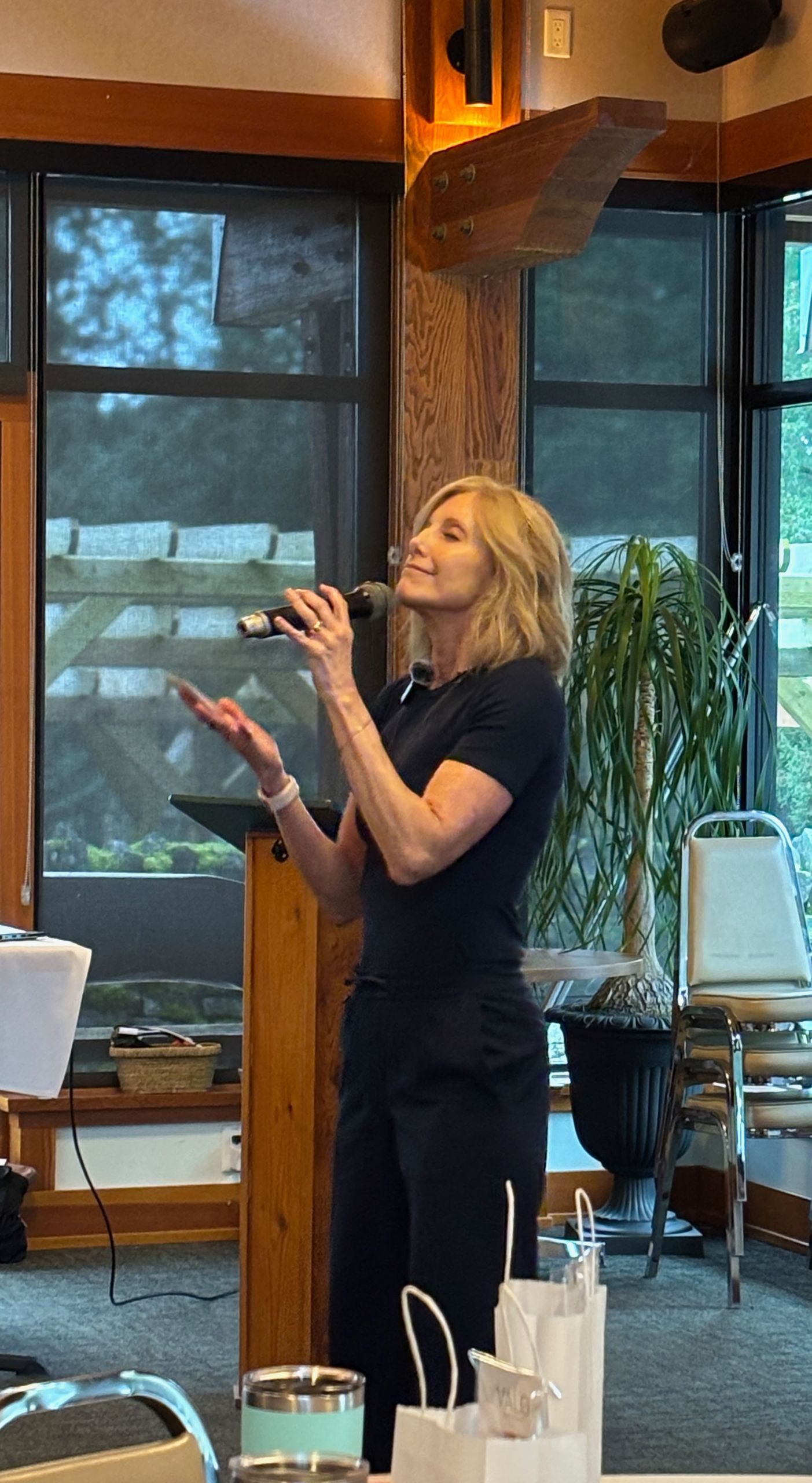 Woman with blonde hair speaks into a microphone at a podium. She is dressed in black with a slight smile, gesturing in a wood-paneled room with large windows.