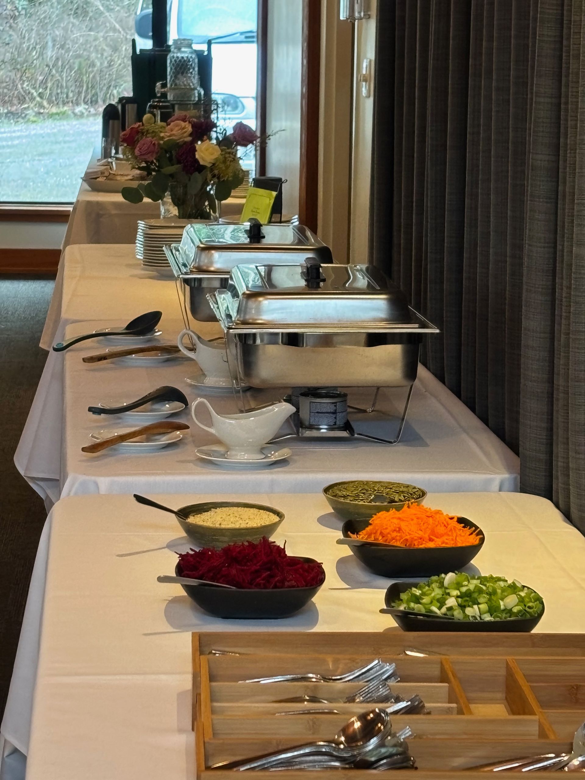 Buffet table with chafing dishes, bowls of vegetables, and silverware. Flowers and windows are also in the background.