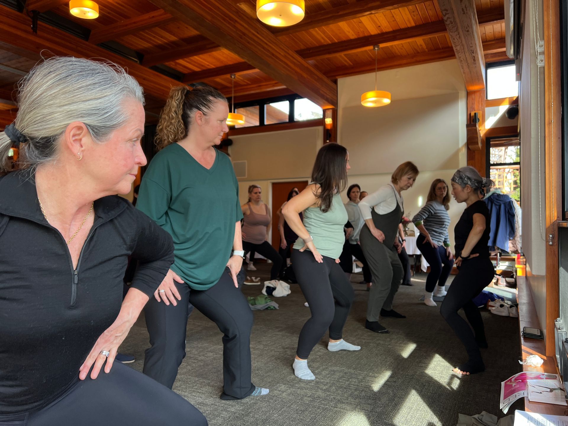 A group of women doing yoga or dance moves in a wood-paneled room with natural light. They wear workout clothes and have varying skin tones.