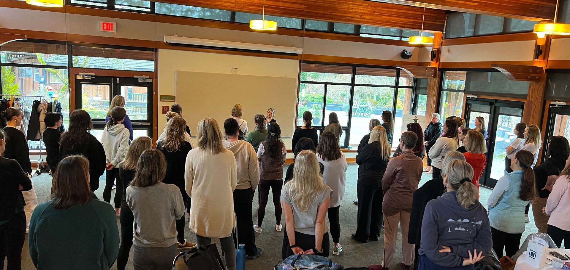A group of people standing in a large room with large windows, some facing a person at the front.