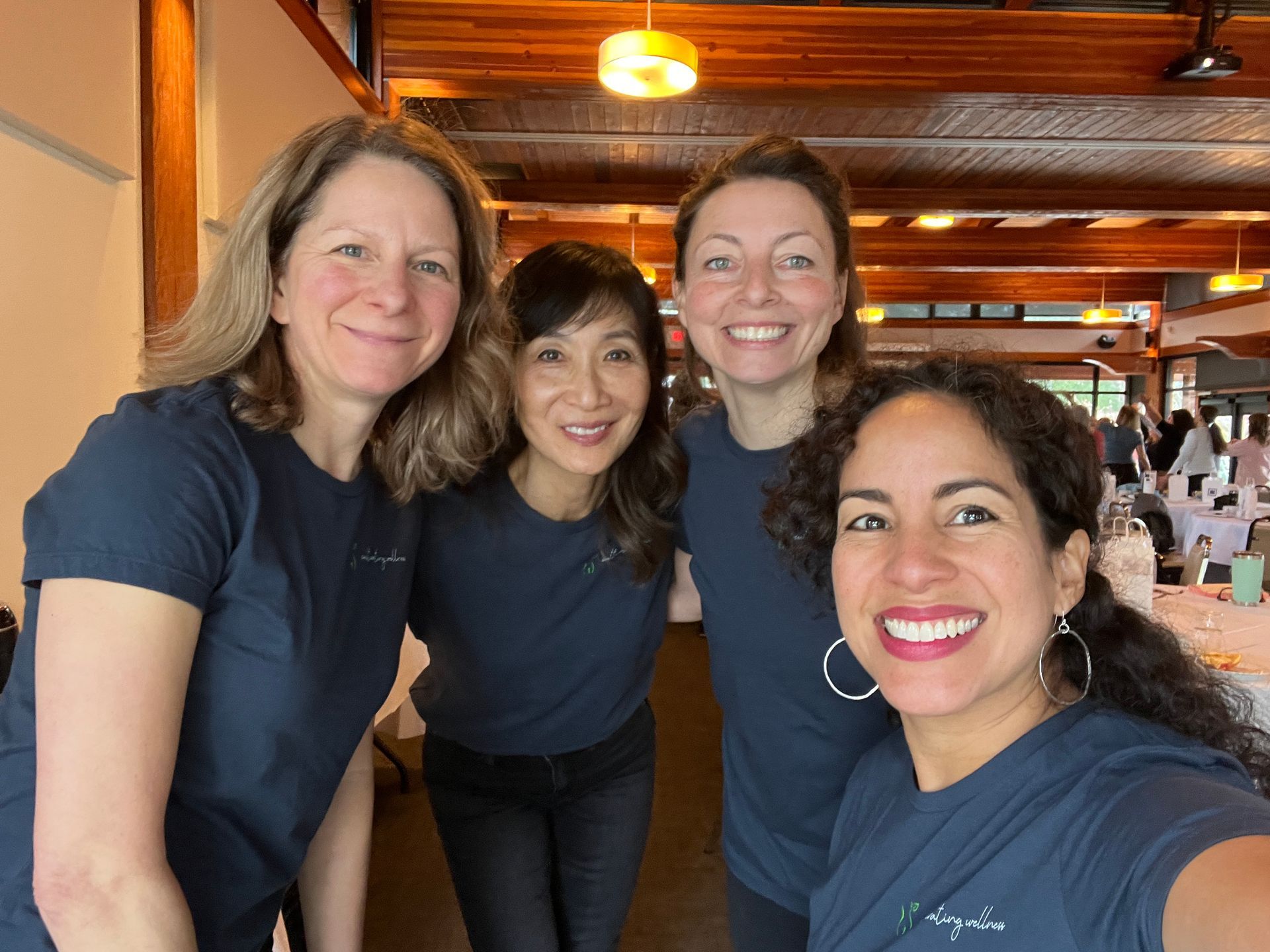 Four women wearing matching blue shirts smile together indoors. They're in a brightly lit room with a wooden ceiling and some guests in the background.