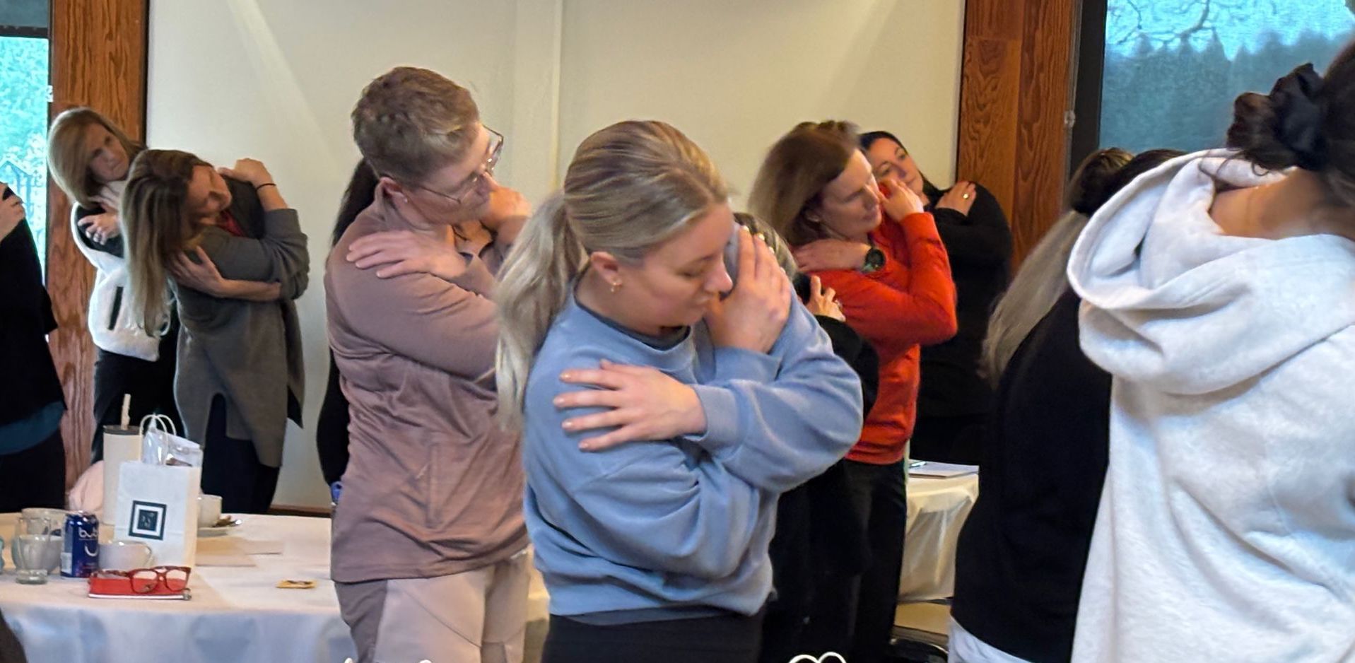 A group of women in a well-lit room performing a self-hugging exercise with eyes closed. They are likely participating in a wellness workshop.