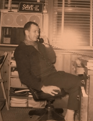 Man on phone, seated in office. Sepia tone. Air conditioner above, blinds, paper stack and desk.