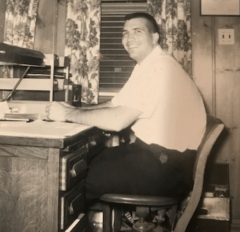 Man in uniform smiles at a desk, seated in a wood-paneled room, near a window.